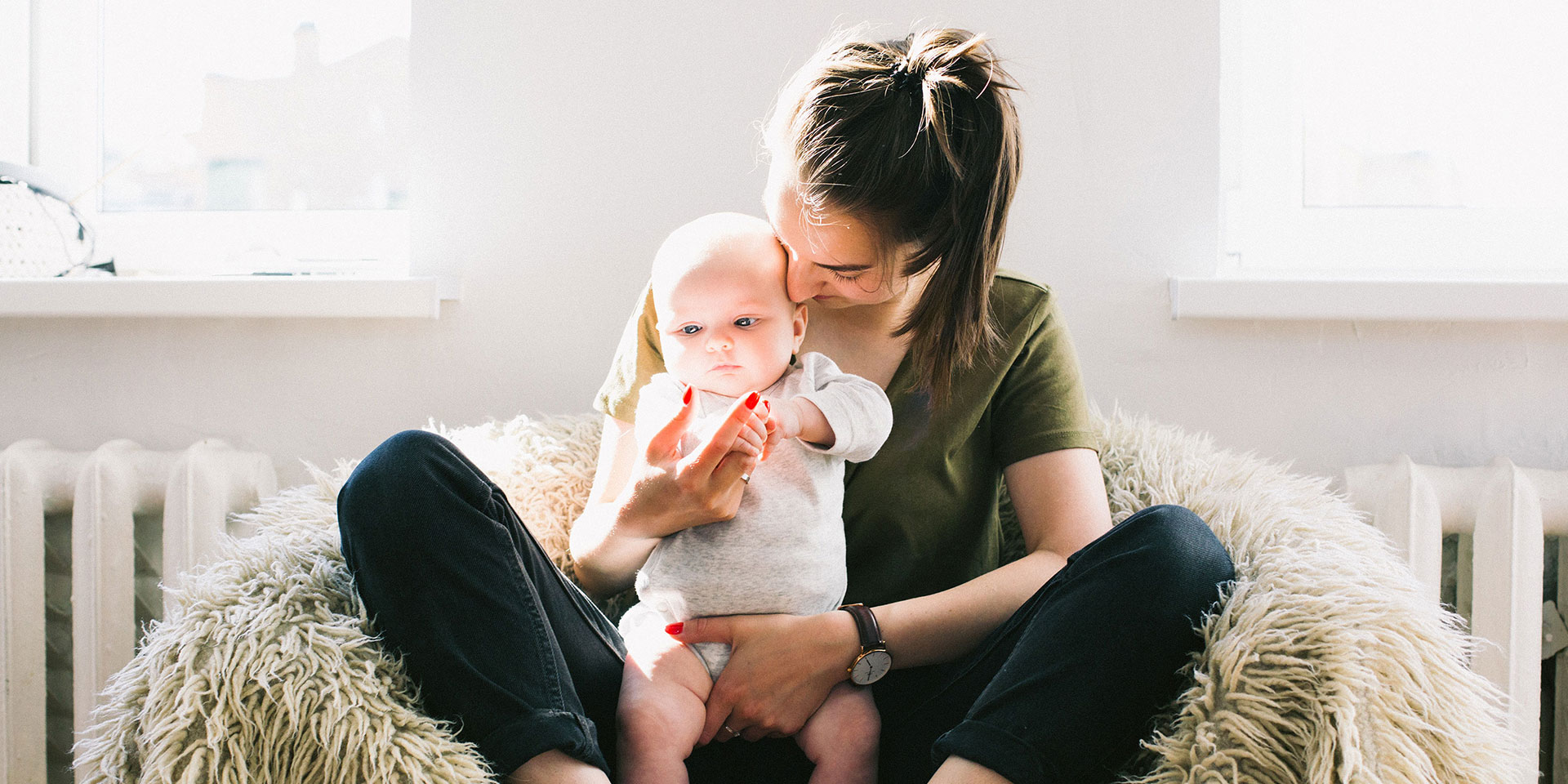 A woman sitting down on a soft puff, holding a standing baby.